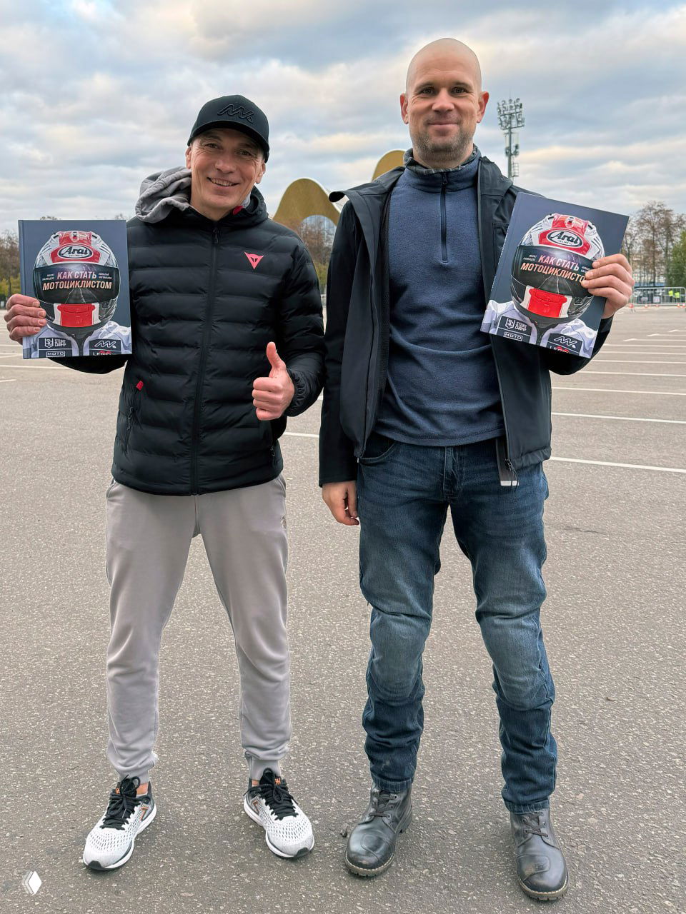 Два автора стоят на открытой парковке и держат по экземпляру книги «Как стать мотоциклистом», на обложке — шлем и крупный логотип.