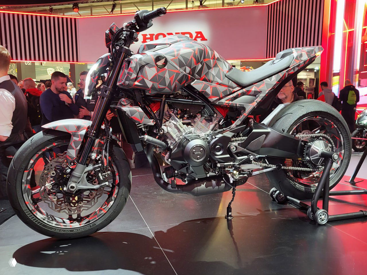 Прототип нейкеда Honda на выставке EICMA в камуфляжной плёнке, открытый корпус и видимый трёхцилиндровый мотор с сильной архитектурой шасси на подставке.