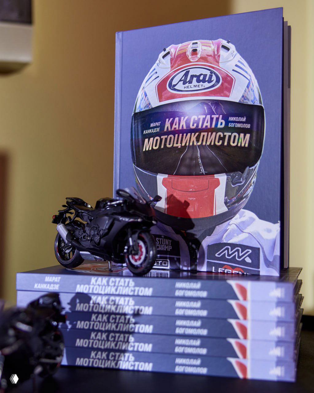 Стопка книг «Как стать мотоциклистом» с обложкой-шлемом; на переднем плане — миниатюрная модель спортивного мотоцикла.
