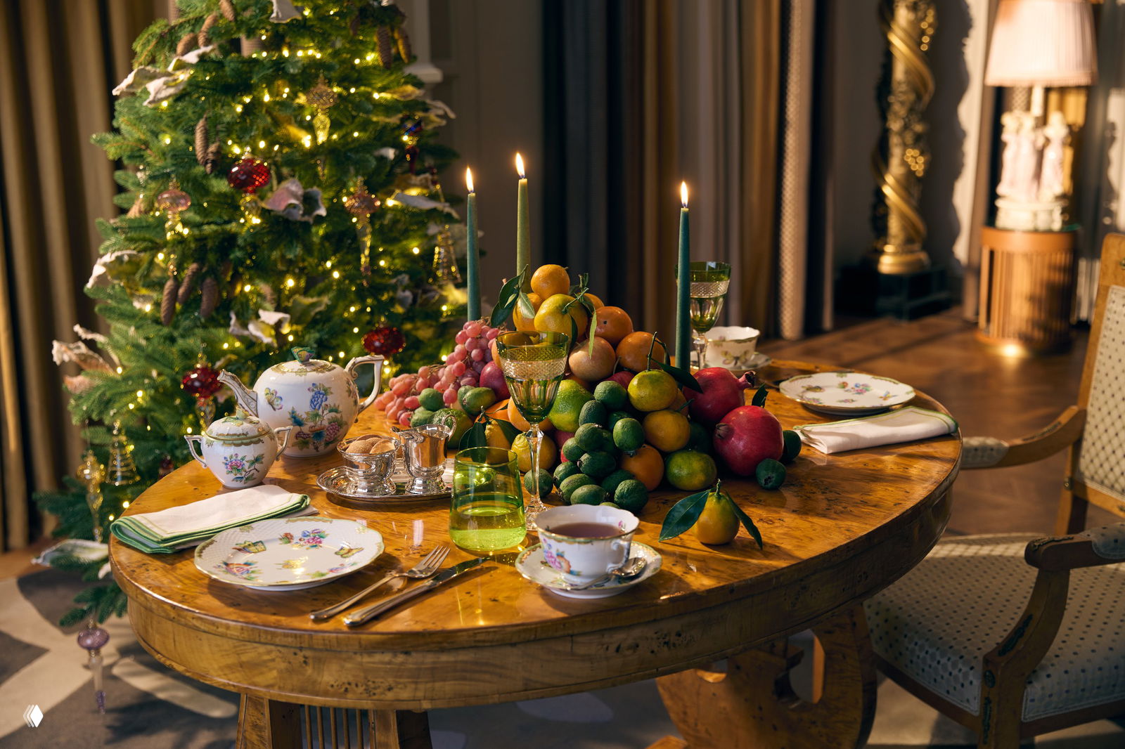 Деревянный стол с фруктовой композицией, фарфоровым сервизом и свечами рядом с украшенной рождественской ёлкой.