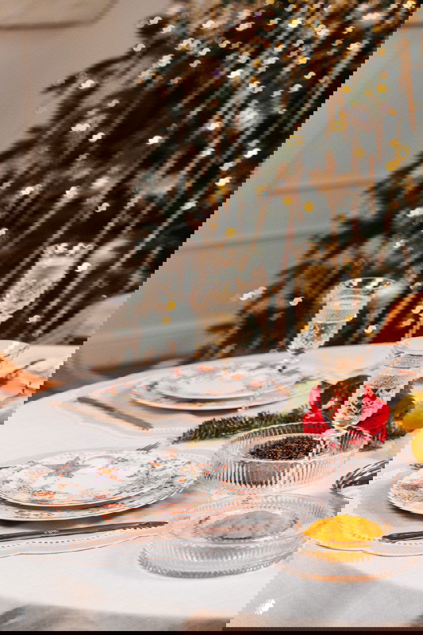 Горизонтальное фото: праздничный стол с сервировкой из коллекции «Счастливая звезда», бокалами и размытой ёлкой на заднем плане для атмосферного кадра.