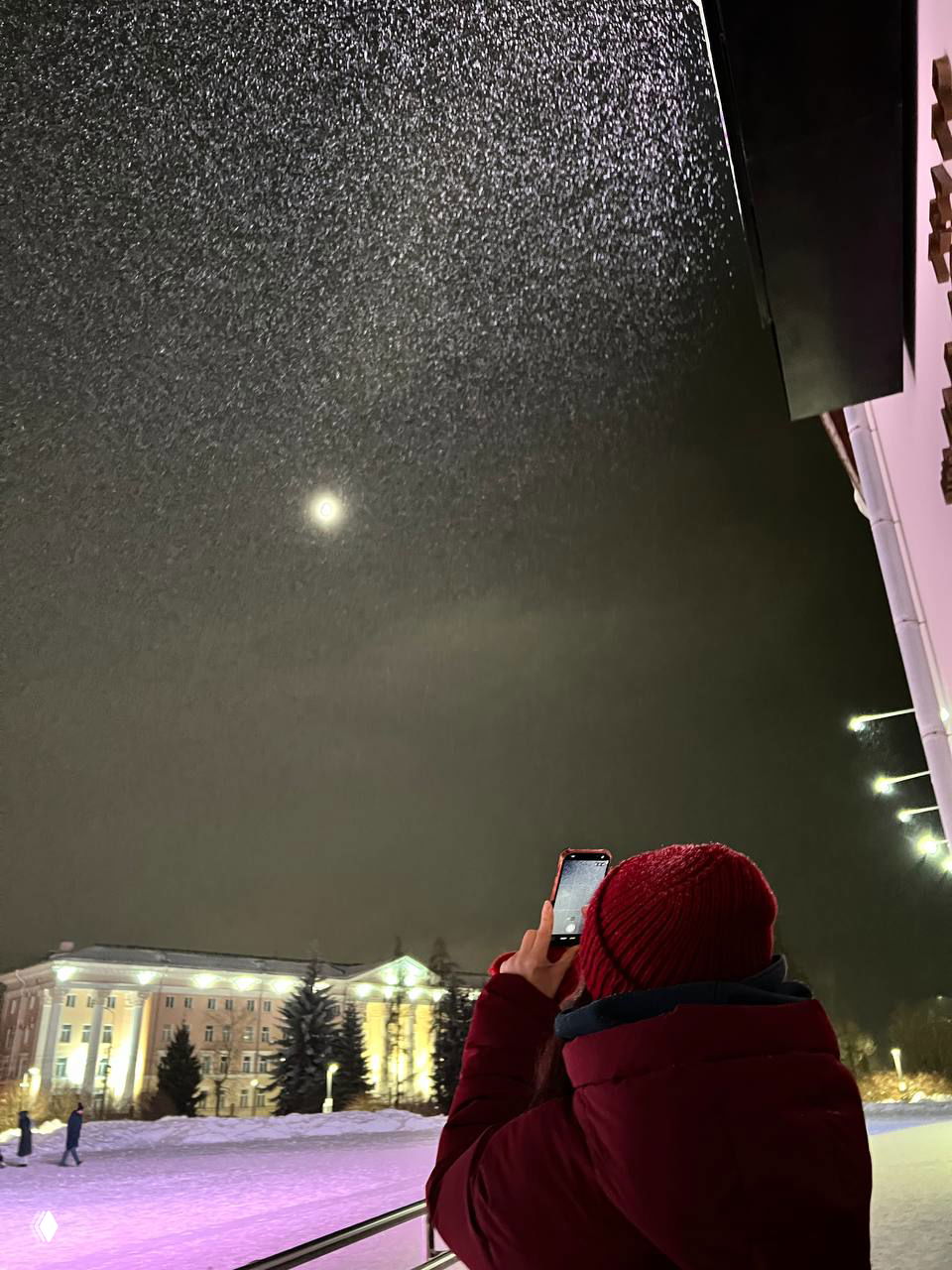 Человек в тёплой красной куртке и шапке фотографирует ночное небо с луной и падающим снегом на освещённой городской площади у зданий.