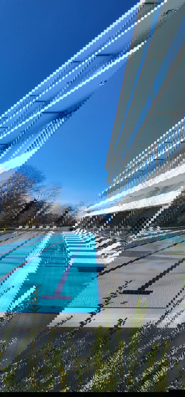 Бассейн у фасада современного отеля: дорожки для плавания, шезлонги на террасе и аккуратная озеленённая зона под ясным небом.