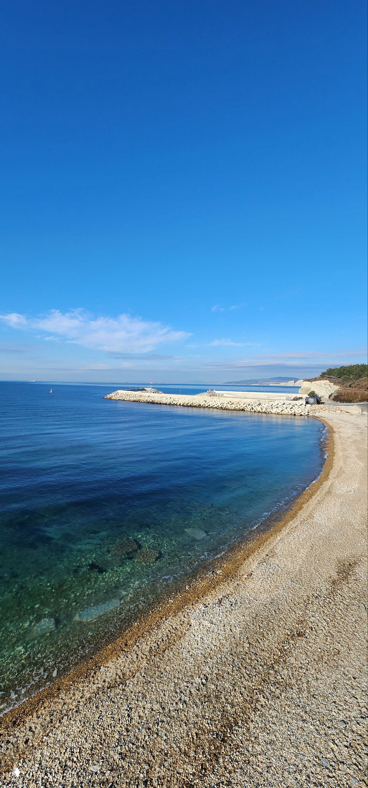 Панорамный вид на море и гальку побережья у Геленджика: спокойная бирюзовая вода, волнорез и широкая полоса чистого неба.