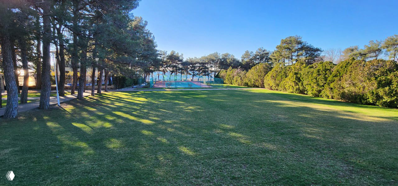 Озеленённая территория санатория: аккуратно подстриженный газон и теннисная площадка вдали, просторная зона для прогулок и активности на свежем воздухе.