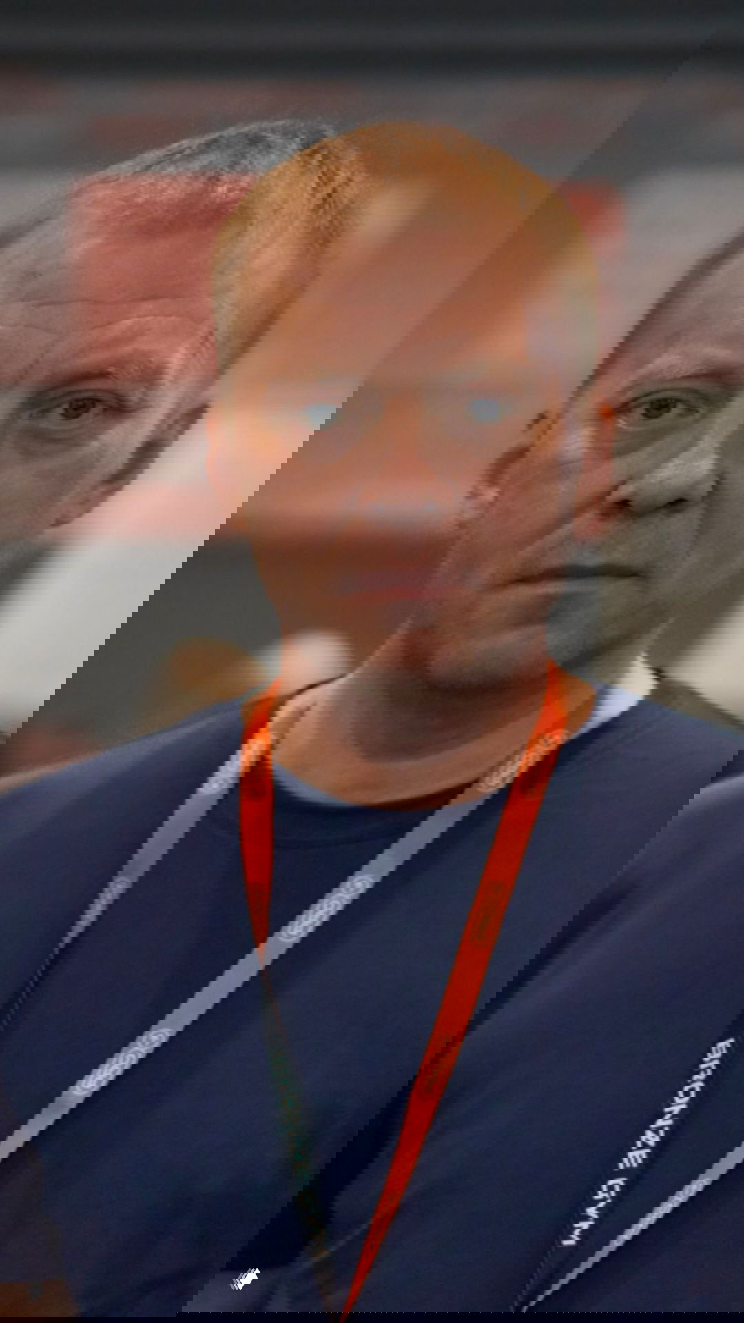 Крупный план мужчины с бейджем на оранжевом шнурке; нейтральное выражение лица и размытый фон конференции.