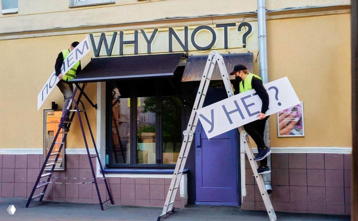 Двое рабочих на лестницах меняют англоязычную вывеску «WHY NOT?» на русский вариант у фасада небольшого магазина в городе.
