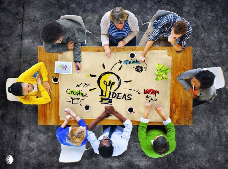 Вид сверху на команду за столом с рисунком лампочки и надписью IDEAS — мозговой штурм идей для салона и контента.