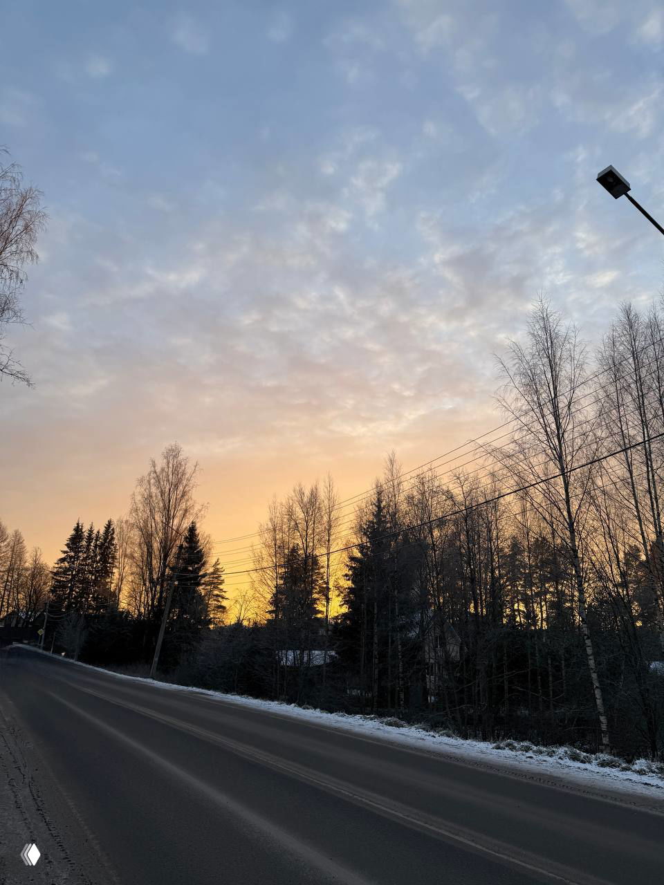 Пустая просёлочная дорога на фоне заката, силуэты берёз и сосен и мягкое оранжево‑голубое небо над загородной местностью рядом с домом.