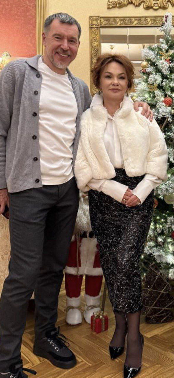 Два человека в вечерней одежде позируют у украшенной рождественской ёлки в интерьере театра; праздничная обстановка и декор.