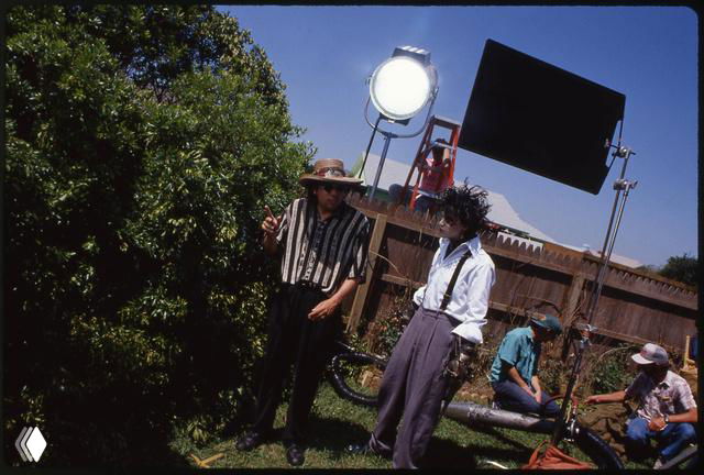 Внешняя съёмочная площадка: персонаж в костюме стоит под дневным светом на фоне техники и членов команды, видно осветительные рамы.