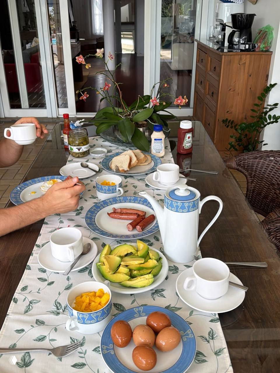 Завтрак на столе: авокадо, яйца, сосиски, чашки, хлеб и соусы на сервированной столешнице в домашней столовой с цветами в центре.