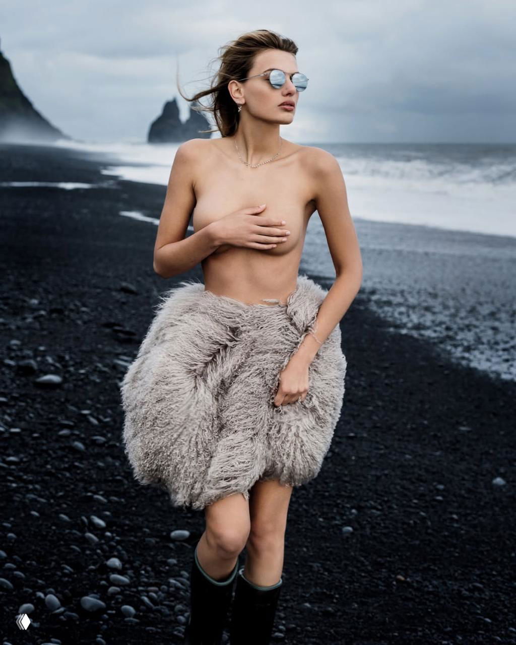Модель на чёрном пляже в полупрозрачной шубке и сапогах; поза с прикрытой рукой, драматичный морской пейзаж подчёркивает нордическую эстетику съёмки.