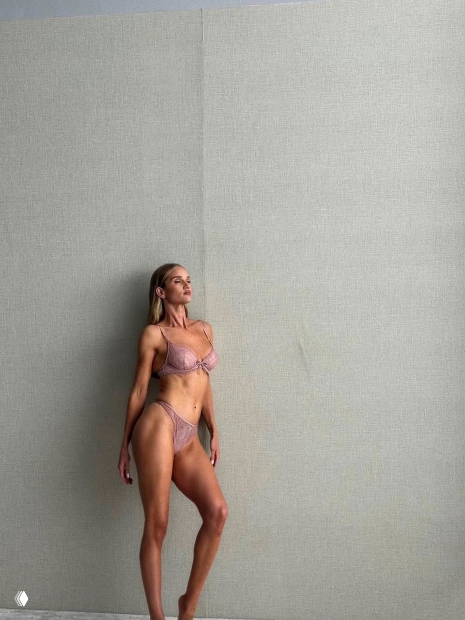 Модель в кружевном розовом белье на фоне светлой фактурной стены; высокий ростовой кадр, изящная поза стоя в студии.