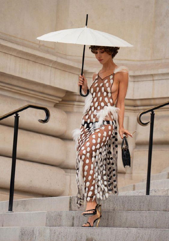 Модель спускается по ступеням в прозрачном платье с белыми элементами, держит белый зонтик и маленькую сумку.