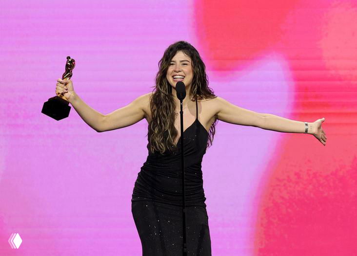 Женщина в чёрном платье держит статуэтку AVN Awards и благодарит публику за победу на сцене с розово-белым фоном и микрофоном.