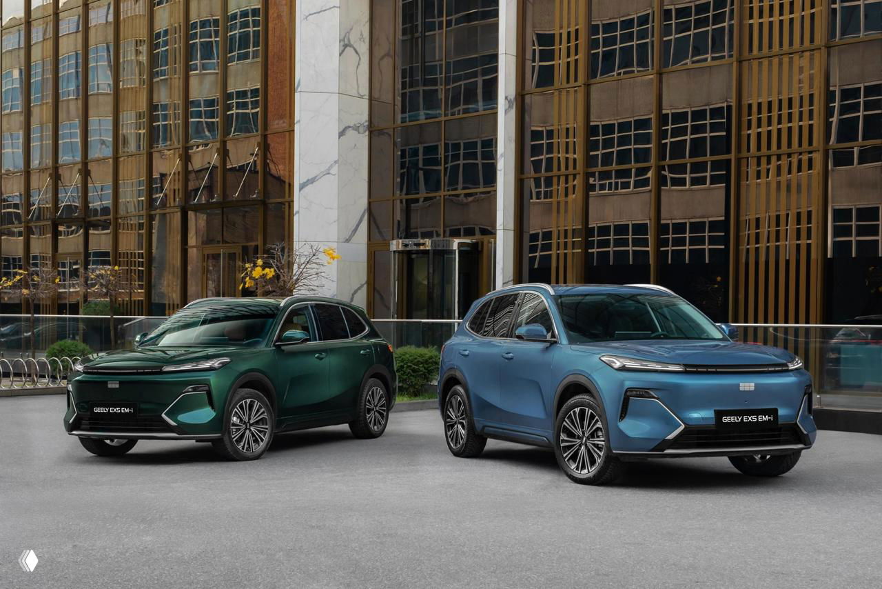 Два кроссовера Geely EX5 EM-i (синий и зелёный) на стоянке у современного офисного здания — общий план фасада и передней части автомобилей.