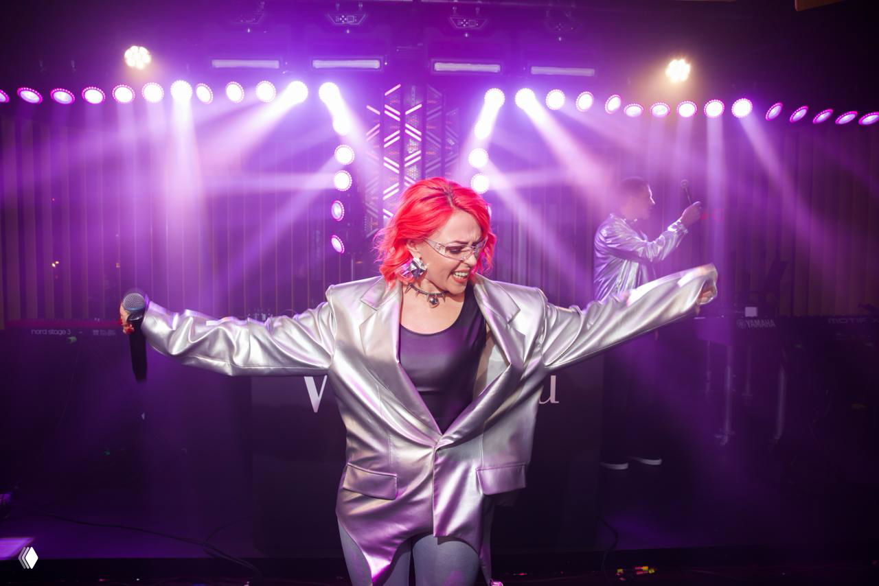 Певица в блестящем серебристом пиджаке выступает на сцене с микрофоном, вокруг заметны пурпурные прожекторы и атмосферный световой дым