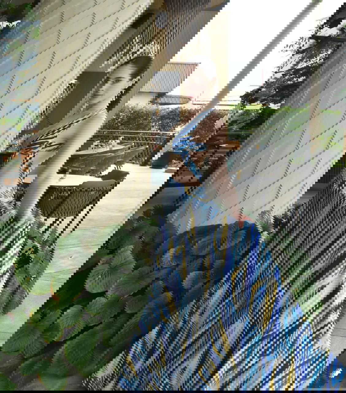 Женщина в длинном платье с синим узором у колонны и зелёных растений — поза в полный рост в зоне ресторана или лаунжа у бассейна.