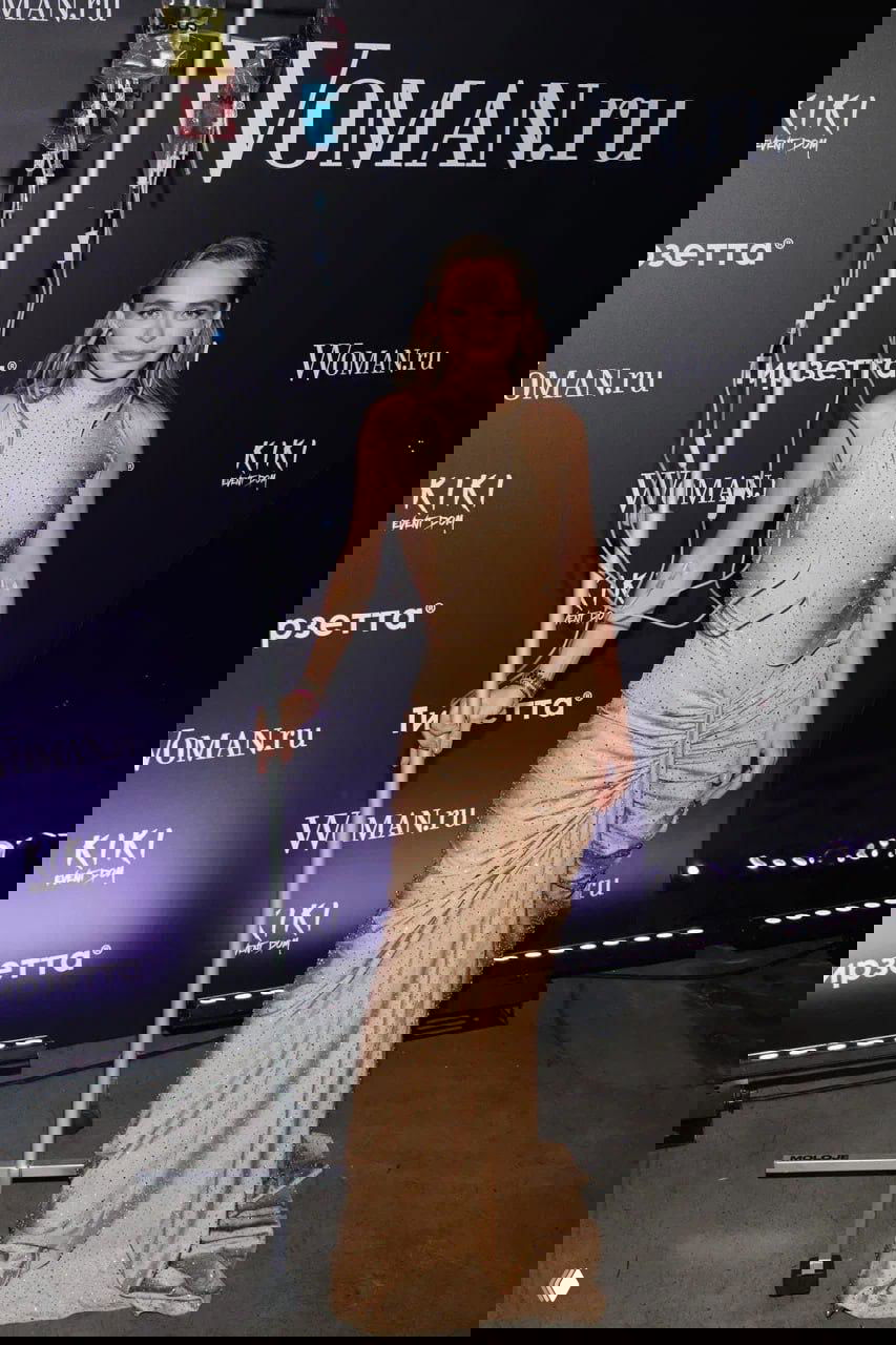 Женщина в нюдово-золотистом платье на красной дорожке рядом с брендовой стенной с логотипом Woman.ru и фиолетовой подсветкой зоны фотозоны