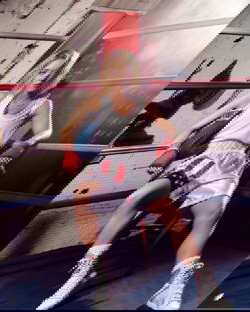 Сидни Суини сидит в ринге в шортах с американским флагом и боксерской перчаткой рядом, дымчатая атмосфера и постановочный свет.