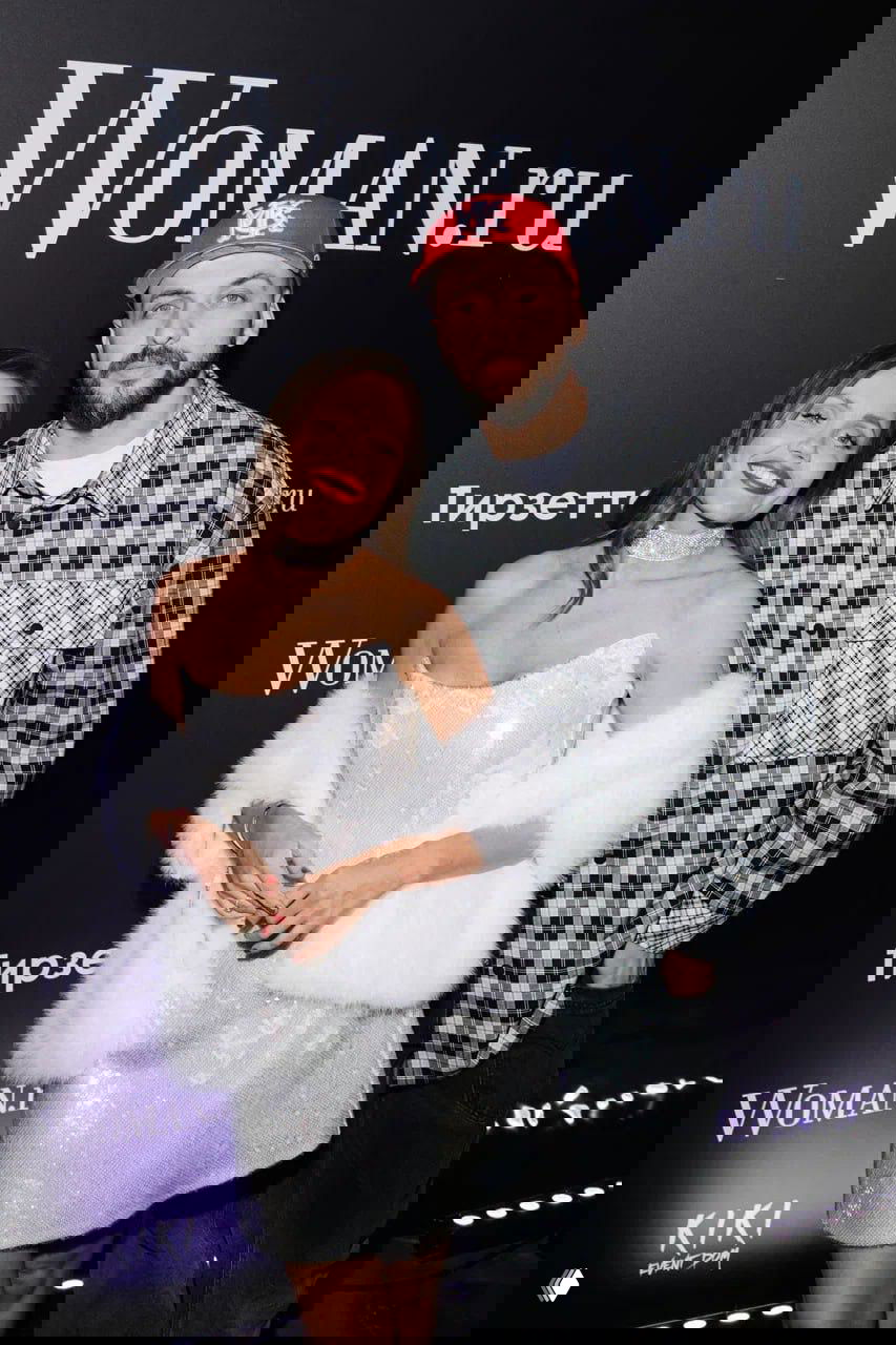 Пара на фоне брендированного баннера стоит в неформальном образе у фотозоны, вокруг заметна логистика мероприятия и фиолетовая подсветка