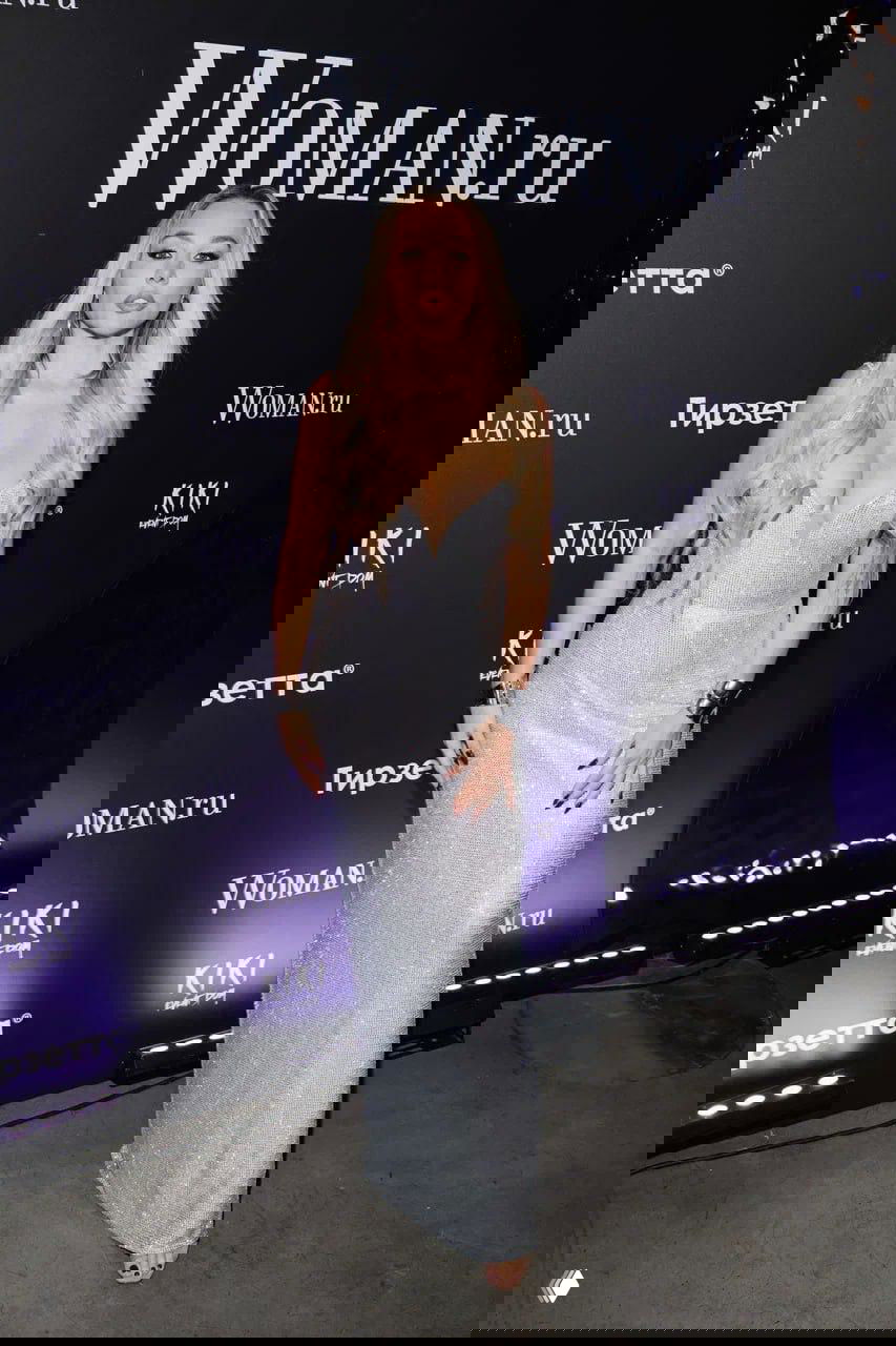 Женщина в длинном серебристом платье позирует у стенда Woman.ru на красной дорожке мероприятия с эффектным световым контрастом