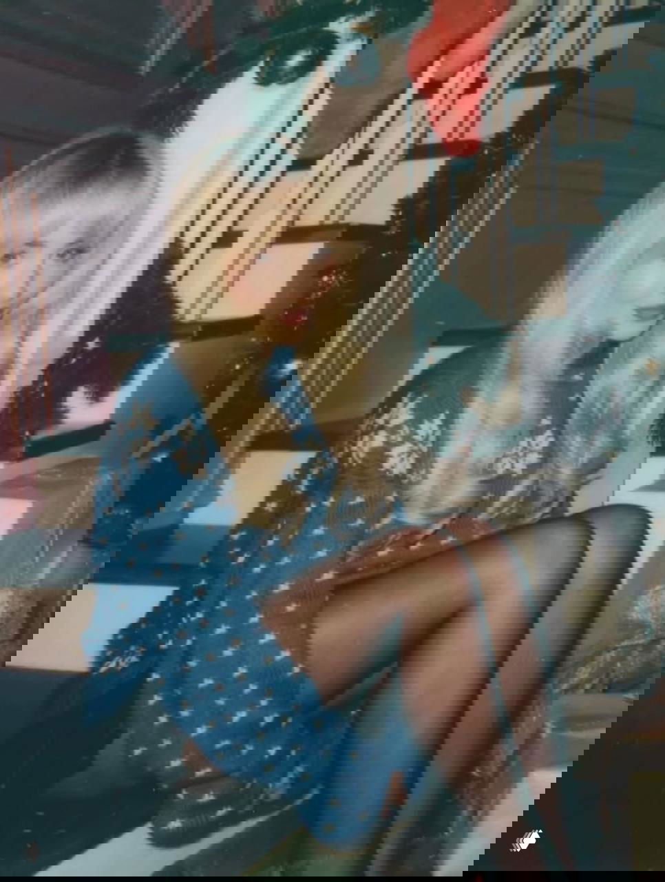 Женщина в тёплом свитере и чулках сидит на ступеньках рядом с ёлкой и подарками, кадр подчёркивает праздничное настроение и стиль образа.