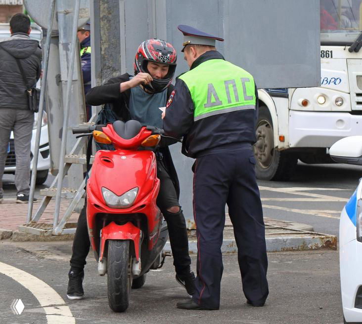 Сотрудник ДПС беседует с водителем скутера у обочины городской дороги на фоне автобуса и припаркованных автомобилей.