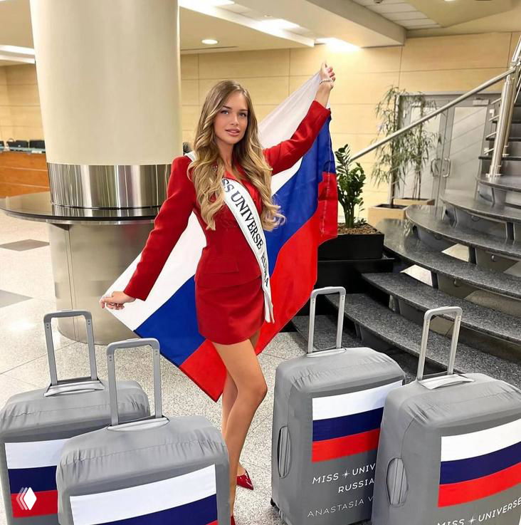 Участница конкурса Miss Universe на фоне большого российского флага и чемоданов с национальными наклейками, кадр из Таиланда