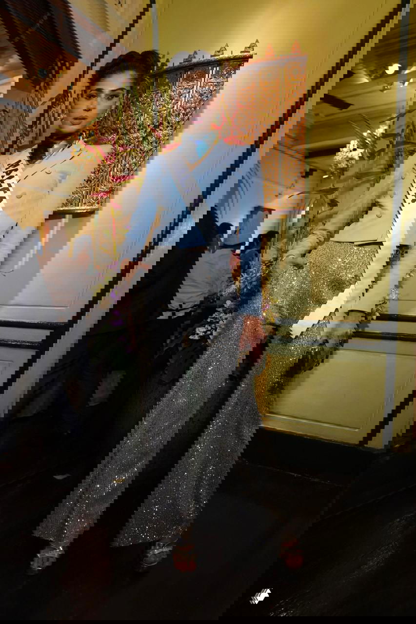 Стройная участница с лентой «MALTA» в стильном наряде и блестящих брюках, фото сделано в помещении перед декором