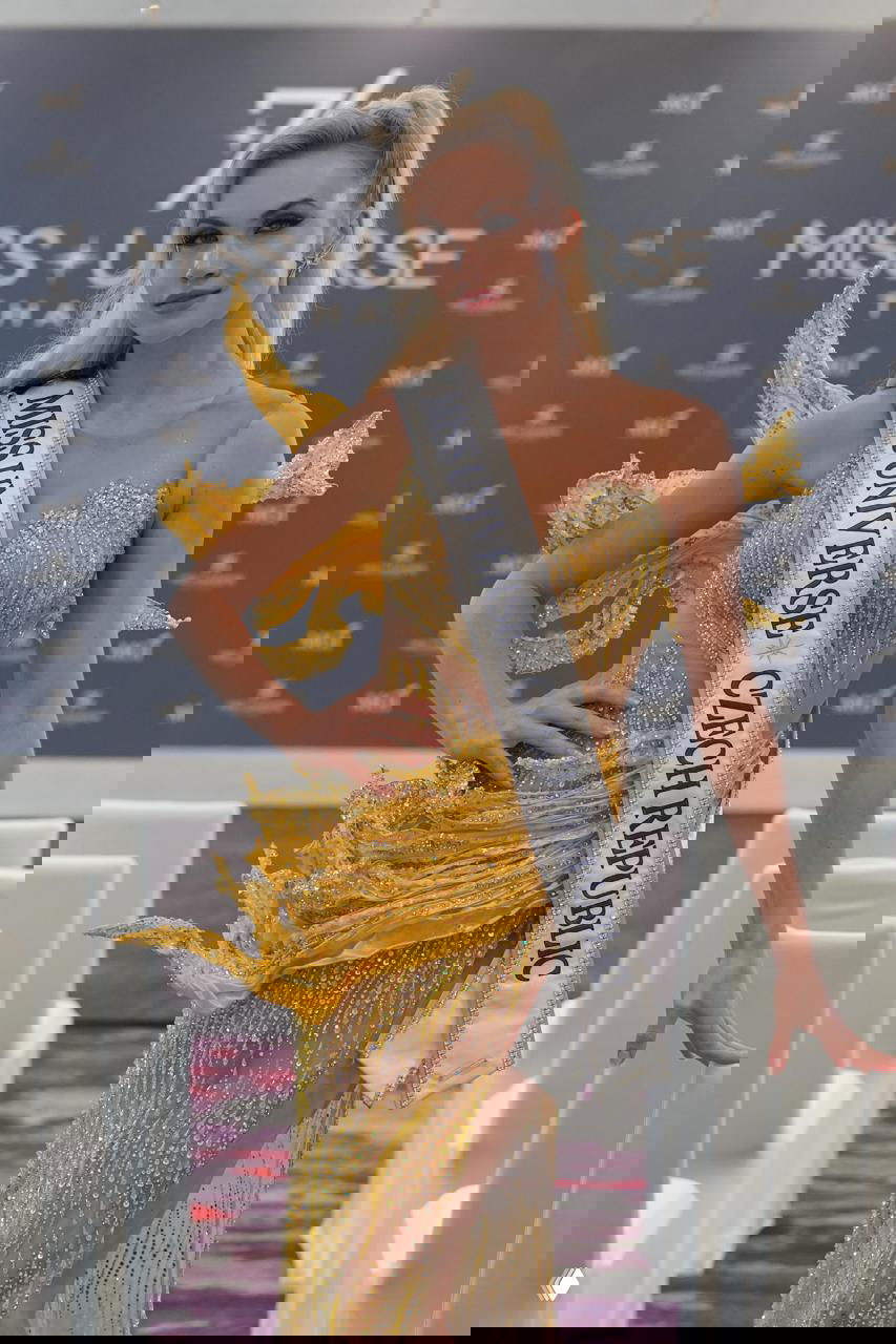 Участница в эффектном золотистом платье с лентой «MISS UNIVERSE CZECH REPUBLIC» на фоне брендированного стенда конкурса