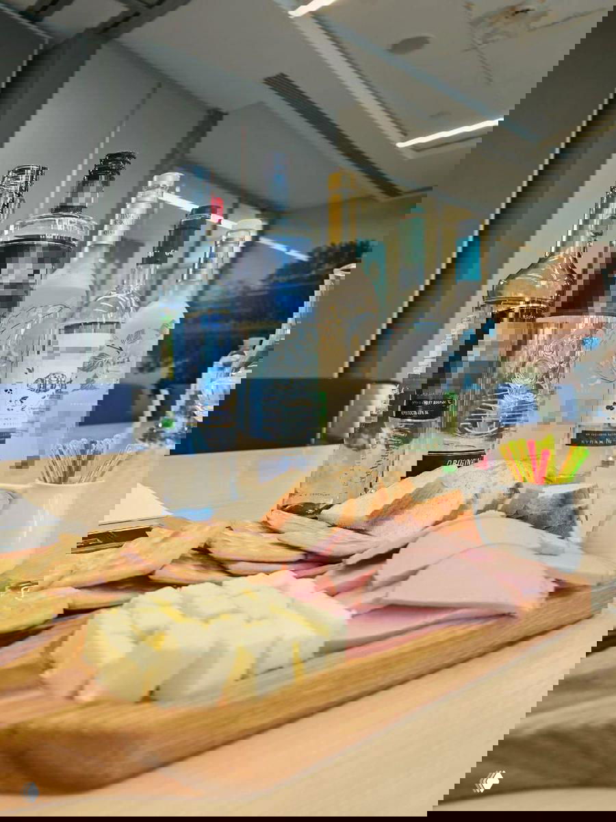 Несколько бутылок водки на столе в офисном интерьере и разделочная доска с хлебом, сыром и колбасой — подготовка дегустации и сопутствующие закуски.
