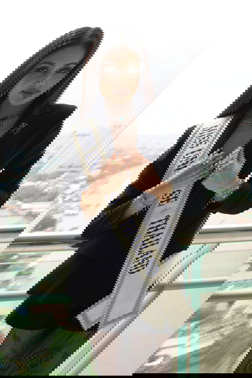 Участница с лентой «MISS UNIVERSE THAILAND» на балконе с видом на город, деловой чёрный костюм и украшения на шее