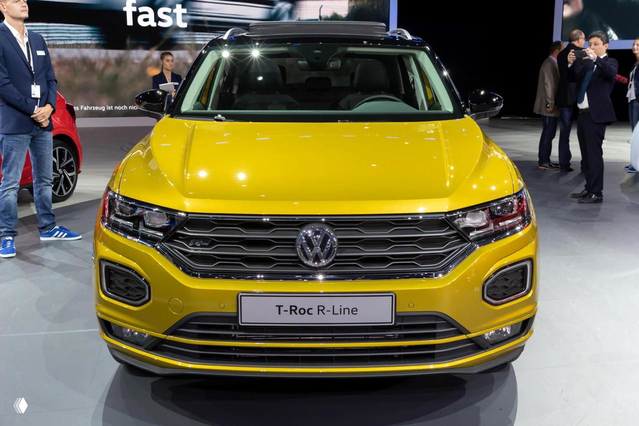 Жёлтый Volkswagen T‑Roc R‑Line спереди на автосалоне; акцент на решётке, бампере и ярком цвете — современный небольшой кроссовер