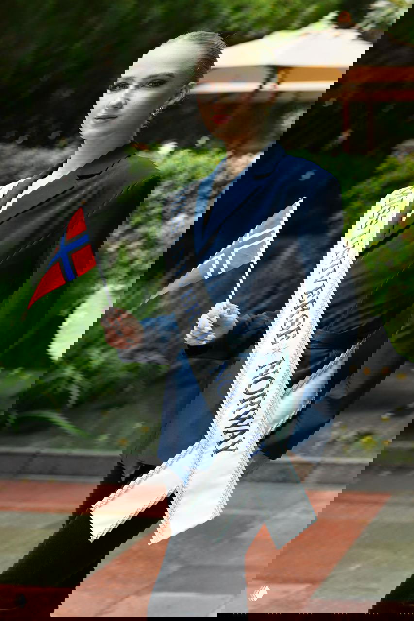 Женщина с лентой «MISS UNIVERSE NORWAY» держит небольшой национальный флаг Норвегии, фотосессия на открытом воздухе