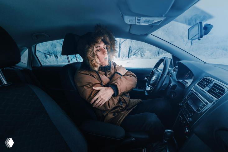 Водитель в зимней куртке сидит в салоне автомобиля с промёрзшими стеклами; кадр иллюстрирует проблему прогрева двигателя и расхода бензина в мороз.