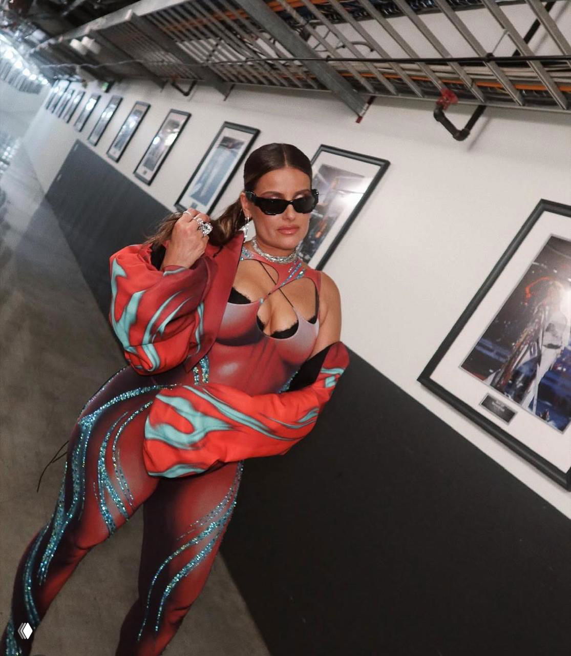 Женщина в ярком сценическом костюме с блёстками и солнцезащитных очках идёт по коридору за кулисами, на стене развешаны фотографии и рамы, полумрак и индустриальные элементы.