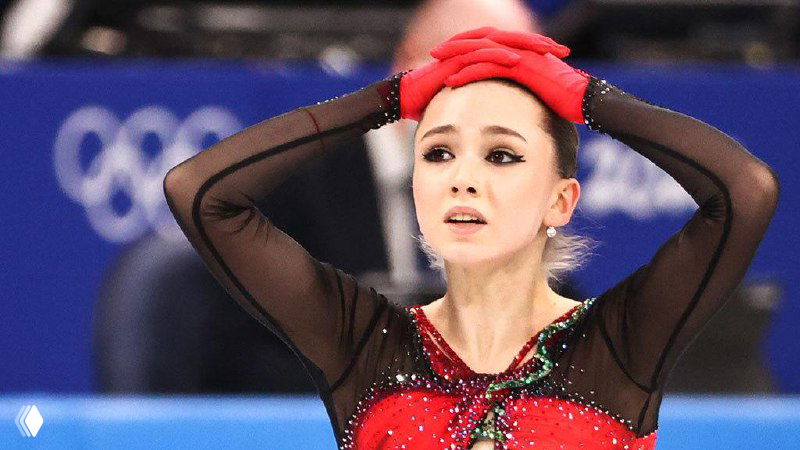 Женщина‑фигуристка в красно‑чёрном костюме на льду, руки на голове и напряжённое выражение; размытый фон с олимпийскими кольцами.