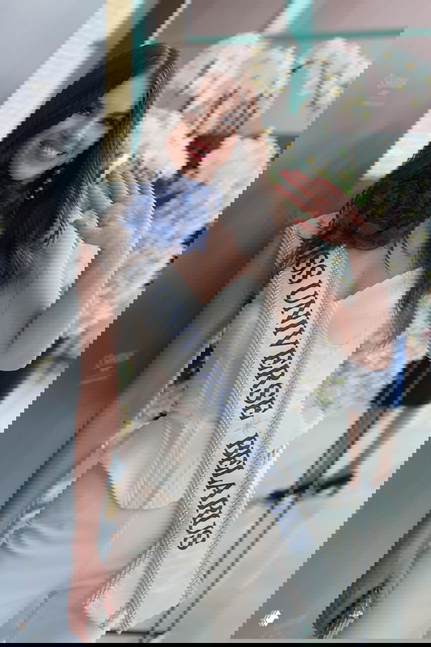 Улыбчивая участница с лентой «MISS UNIVERSE BELARUS» в светлом интерьере отеля, портретный снимок с цветами на фоне