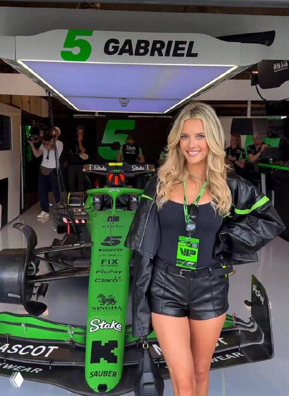 Женщина в чёрной куртке и шортах позирует рядом с зелёным гоночным болидом команды Juncos Hollinger Racing в боксе перед гонкой