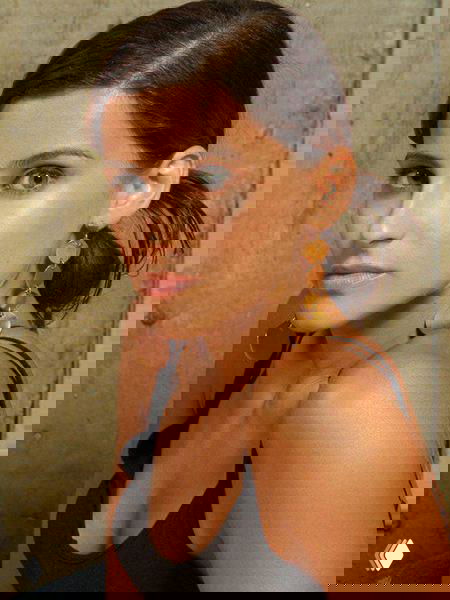 Портрет женщины с зачесанными назад волосами и крупными серьгами‑каплями, нейтральный фон, крупный план лица и плеч, спокойное выражение.