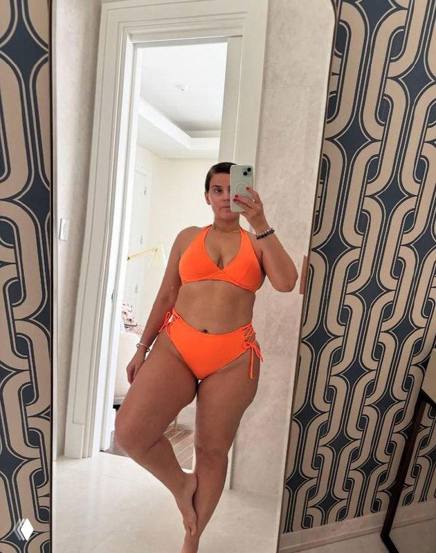 Зеркальное селфи женщины в ярко‑оранжевом бикини в прихожей с узорчатой стеной, полный рост в отражении, в руках виден смартфон и часть интерьера.