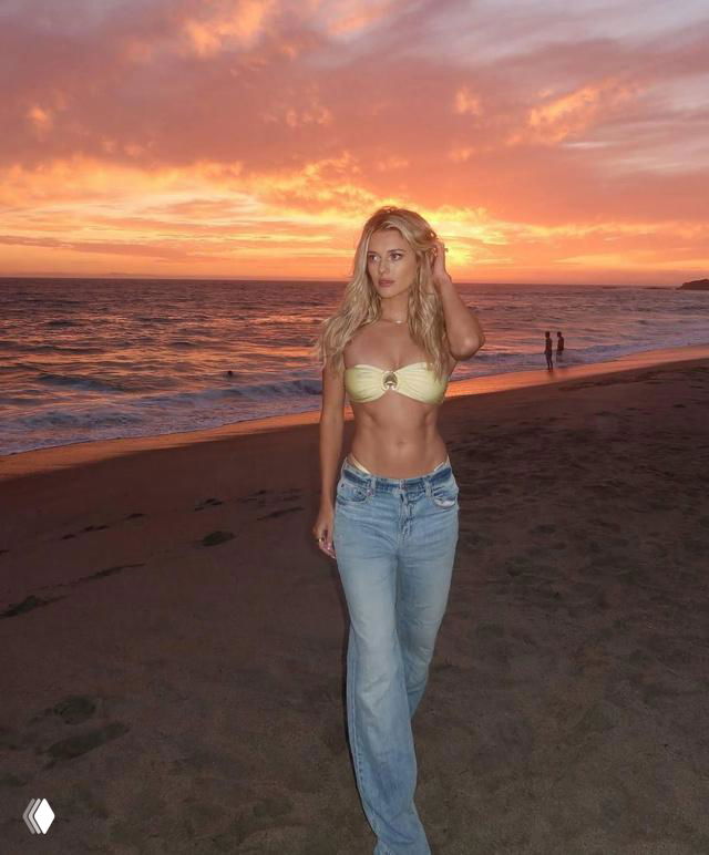 Женщина в купальнике и джинсах стоит на пляже на закате; на горизонте видны море, солнце и силуэты людей вдалеке