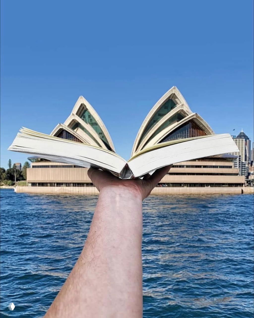 Рука держит раскрытую книгу, страницы которой в кадре формируют контур, напоминающий паруса оперного театра на фоне гавани.