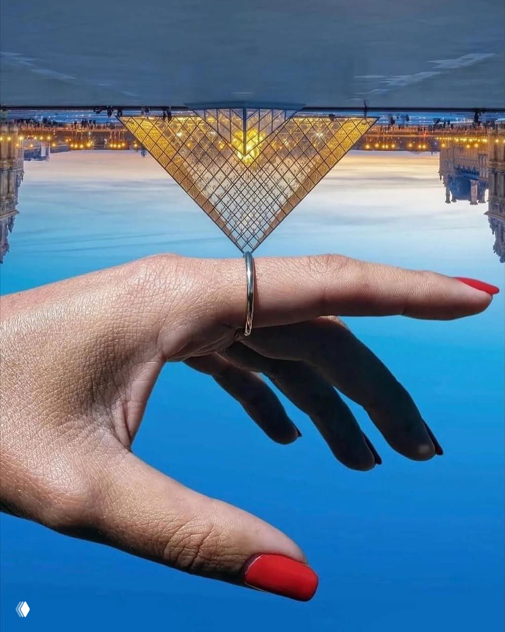 Женская рука с кольцом на пальце на фоне перевёрнутого вида ландшафта; пирамида Лувра визуально «опирается» на палец в зеркальной перспективе.