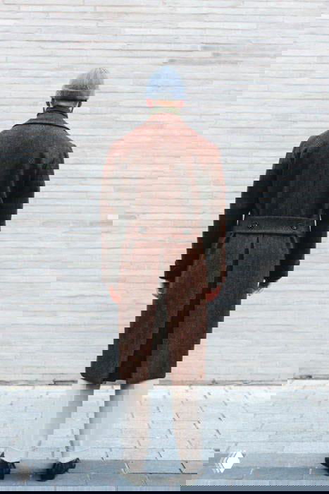 Вид сзади: мужчина в длинном коричневом пальто и серой шапке стоит у светлой кирпичной стены, подчёркнут строгий силуэт и длина изделия.