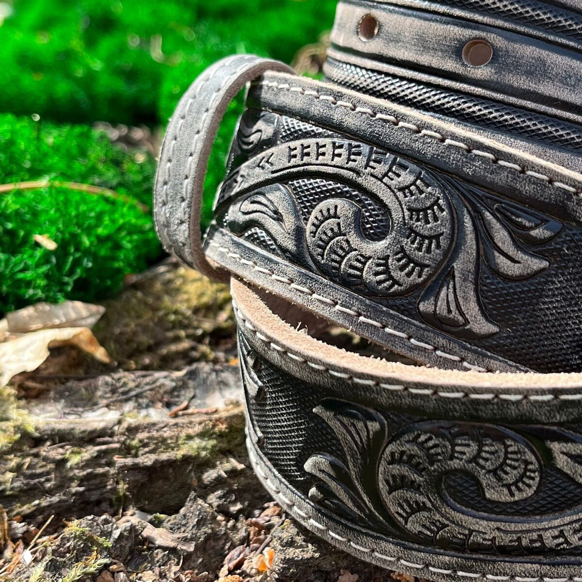 Тёмно-серые кожаные ремни с тонкой строчкой и сложным растительным тиснением, показаны в развёрнутом виде на природном фоне.