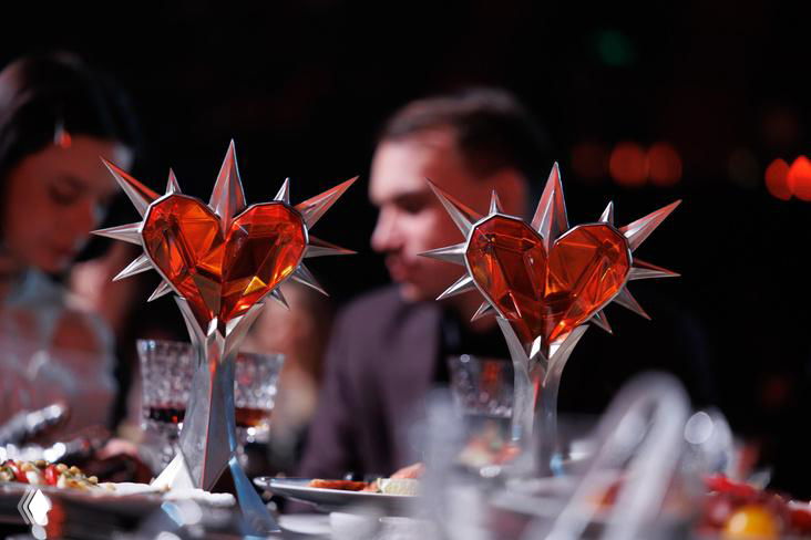 Два красно-кристаллических трофея в форме сердца на банкетном столе церемонии SLAY, на размытом фоне видны гости и сервировка.