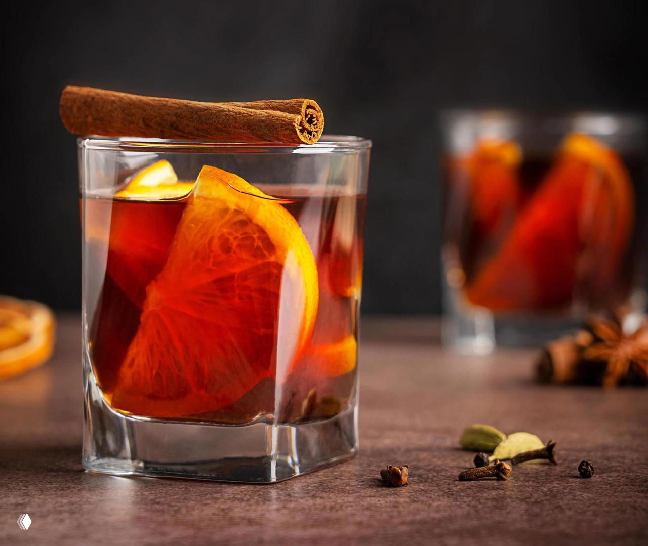 Стакан с тёмной пряной настойкой, долька апельсина и палочка корицы сверху; в кадре целые гвоздики и кардамон.